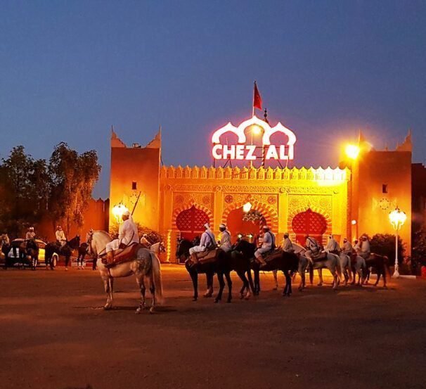 Scène d'un dîner-spectacle au restaurant Chez Ali à Marrakech. La cour est décorée de mosaïques colorées, de lanternes suspendues et de tables recouvertes de tissus somptueux. Des danseuses orientales et des musiciens en tenue traditionnelle animent la soirée, offrant un spectacle vibrant de musique et de danse marocaines. En arrière-plan, une grande tente ornée de rideaux élégants et de coussins crée une ambiance authentique et chaleureuse, typique de l'hospitalité marocaine.